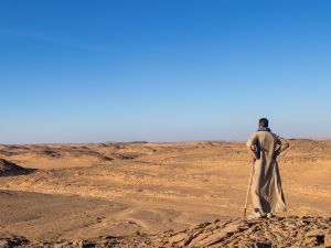 Ein Mann steht auf Wüstenboden und schaut in die Weite der hügeligen Wüstenlandschaft. Der Mann trägt traditionelle arabische Kleidung. Laut Bildbeschriftung wurde das Foto in Ägypten aufgenommen.