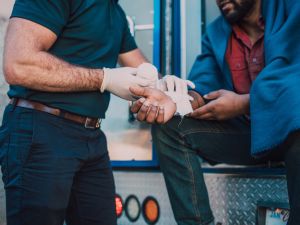 Ein Mann sitzt an der geöffneten rückseitigen Tür eines Fahrzeugs, vielleicht eines Sanitätswagens. Ein Mann mit weißen Handschuhen verbindet das Handgelenk seines rechten Arms mit Verbandszeug.