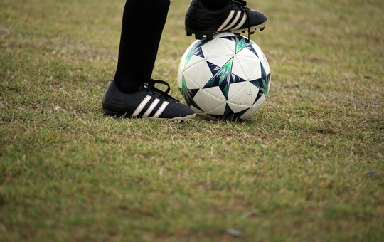 Ein Fußball auf grünem Rasen. Der Ball wird von einem Stollenschuh festgehalten. 