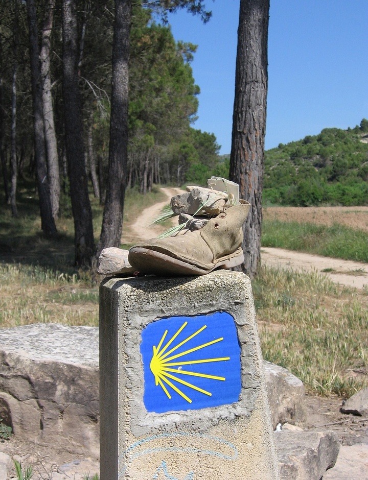 Wanderschuh auf Stein mit gelber Muschel auf blauem Grund für den Jakobsweg