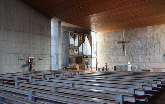 Das Bild zeigt das Innere einer Kirche mit Betonwänden und einer hohen Holzdecke. Reihen von Kirchenbänken stehen vor einem Altarbereich mit einer Orgel und einem Kreuz. Die Beleuchtung ist gedämpft.