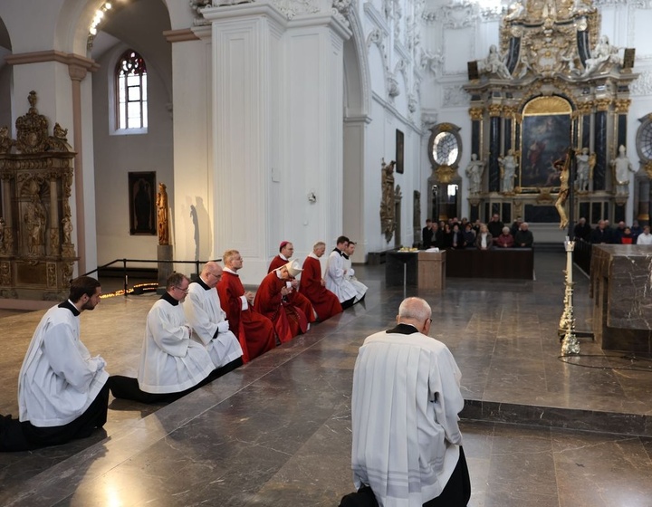 Das Bild zeigt eine kirchliche Zeremonie im Inneren eines prunkvollen Gotteshauses. Mehrere Geistliche in weißen und roten Gewändern knien und sitzen in einer Reihe auf dem Boden. Im Hintergrund sind Kirchenbänke mit Besuchern und ein Altar zu sehen.