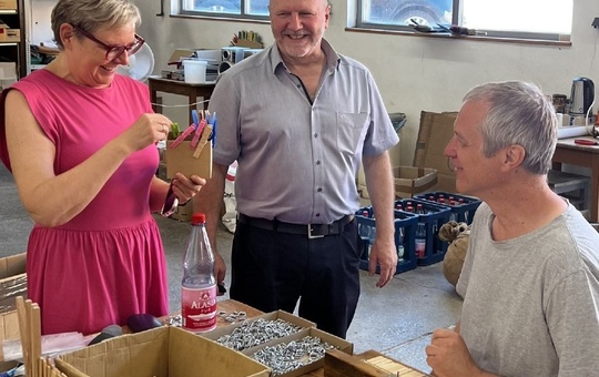 Ein Frau steht mit zwei Männern in einer Werkstatt. Alle drei lachen. Sie hält eine Dose mit Wäscheklammern in der Hand