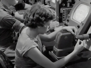 Eine Frau sitzt arbeitend an einer Maschine. Das Foto ist in schwarzweiß.