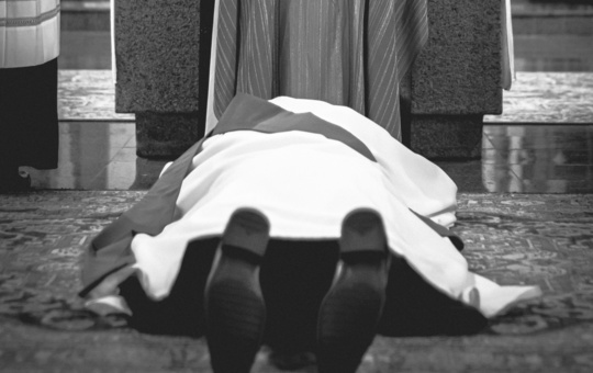 Ein Priester liegt auf dem Boden. Er ist von hinten zu sehen. 