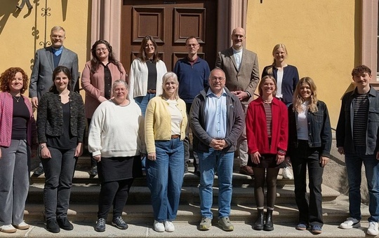 Das Bild zeigt eine Gruppe von Personen, die vor einem gelben Gebäude mit einer braunen Tür stehen. Sie sind auf zwei Stufen verteilt und tragen legere bis formelle Kleidung. Die Gruppe scheint für ein Gruppenfoto zu posieren.