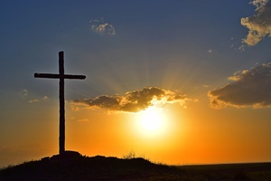 Ein Kreuz steht auf einer Wiese im Sonnenuntergang. Der Himmel ist oben blau und unten gelb.